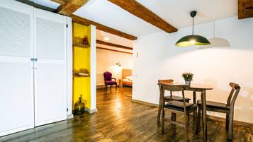 A room with a wooden floor, a dining table with chairs, a yellow wall, and a bed in the background.