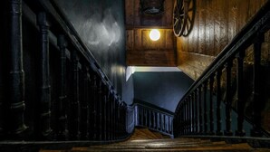 A wooden staircase with a handrail, a light fixture on the wall, and a wheel on the wall. - Blacksmith Old Town Apartments (Tallinn)