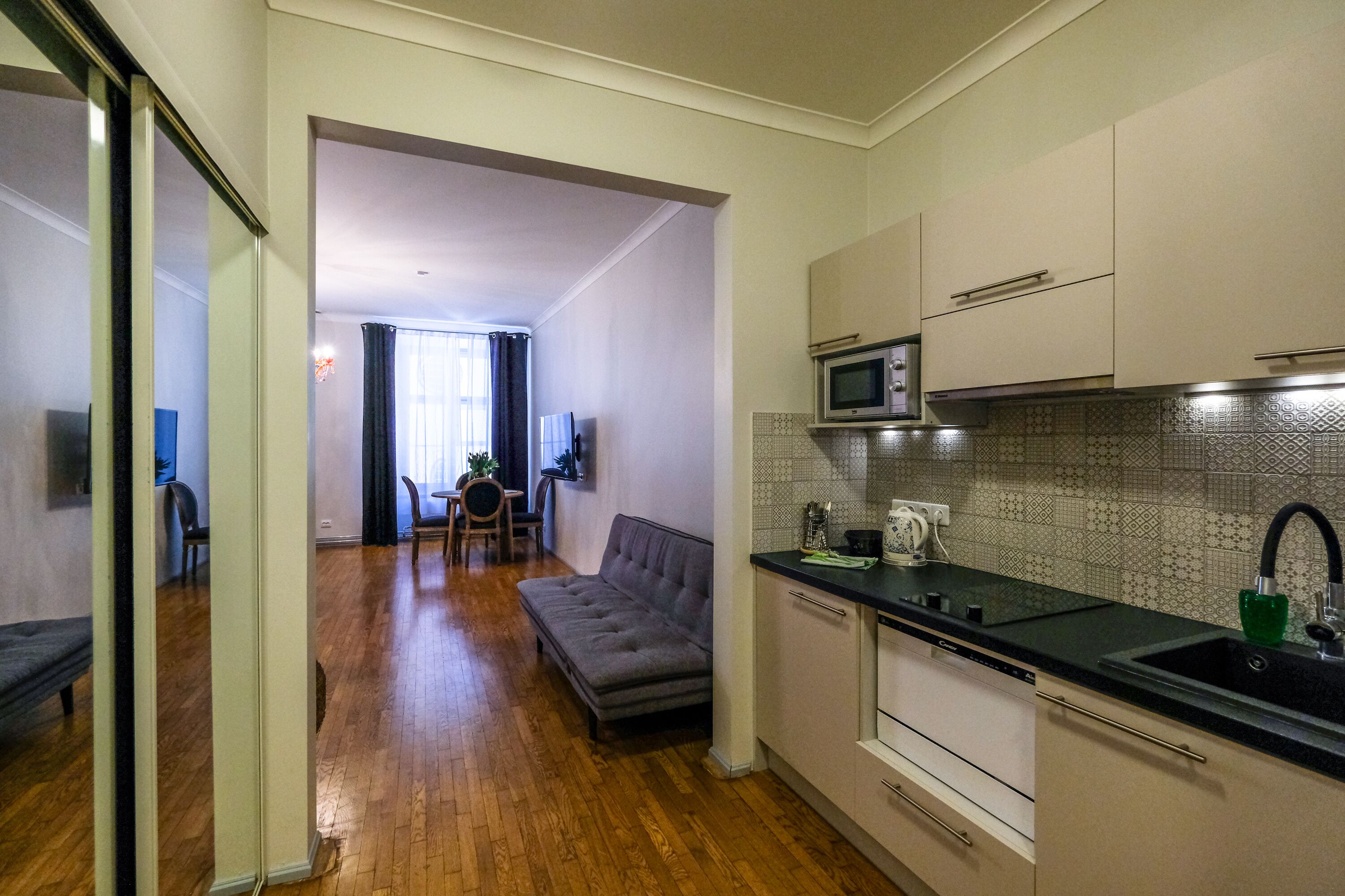 Cocina compacta con microondas, lavabo y muebles de cocina modernos. Un sofá gris se encuentra en la zona de estar, y se ve una mesa de comedor con sillas al fondo.