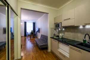 Eine kleine Küche mit Mikrowelle, Spüle und modernen Schränken. Im Wohnbereich steht ein graues Sofa, und im Hintergrund ist ein Esstisch mit Stühlen zu sehen.