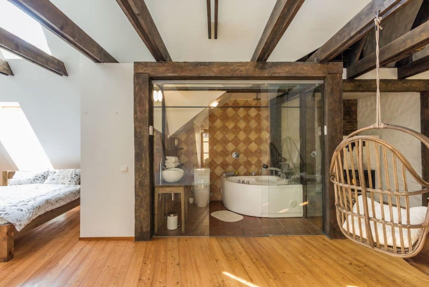 A modern bedroom with a wooden floor, a bed, a hanging chair, and a glass-enclosed bathroom with a bathtub and a tiled wall.