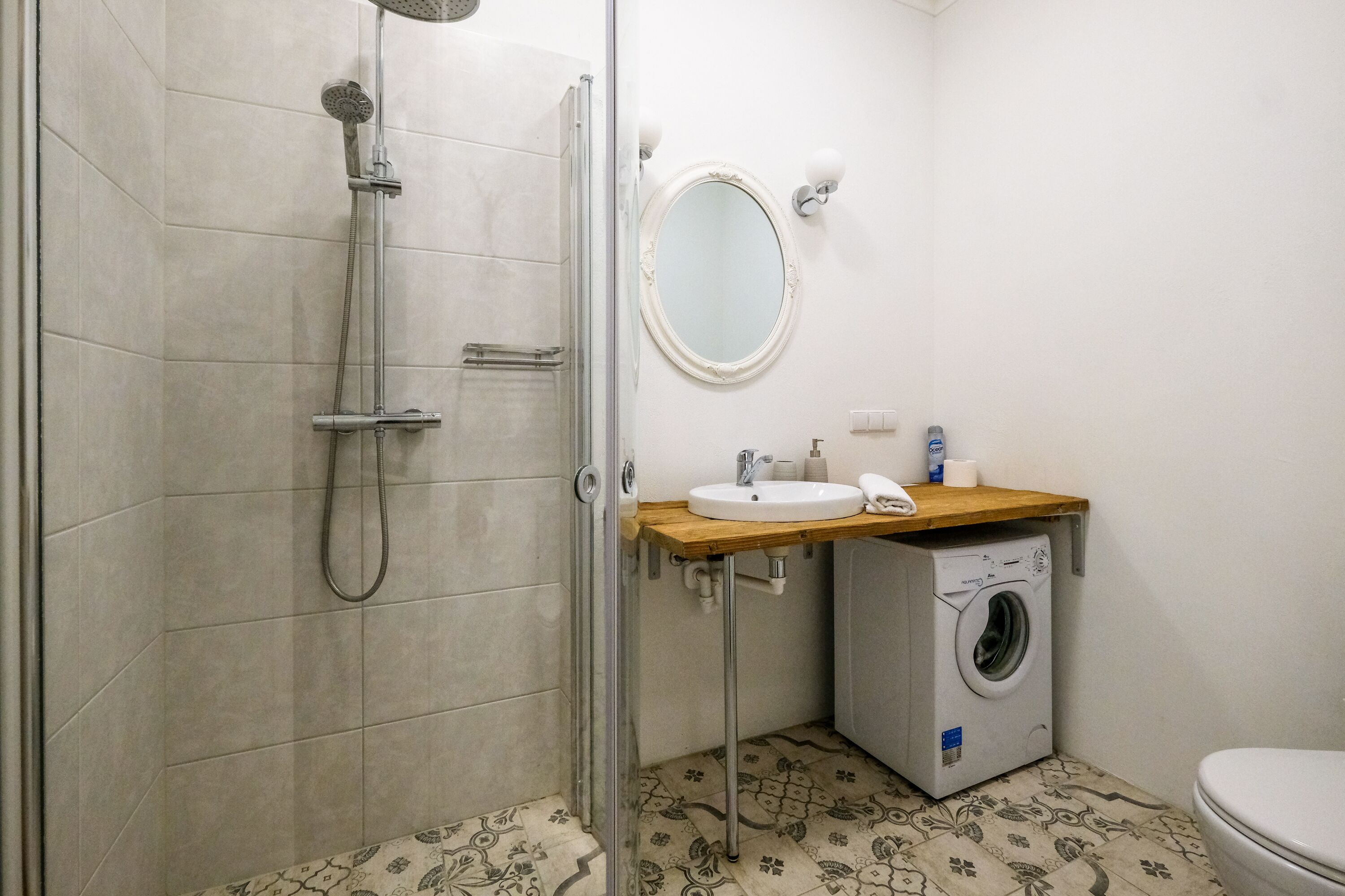 A bathroom with a glass-enclosed shower, a sink with a wooden countertop, a round mirror, and a washing machine.