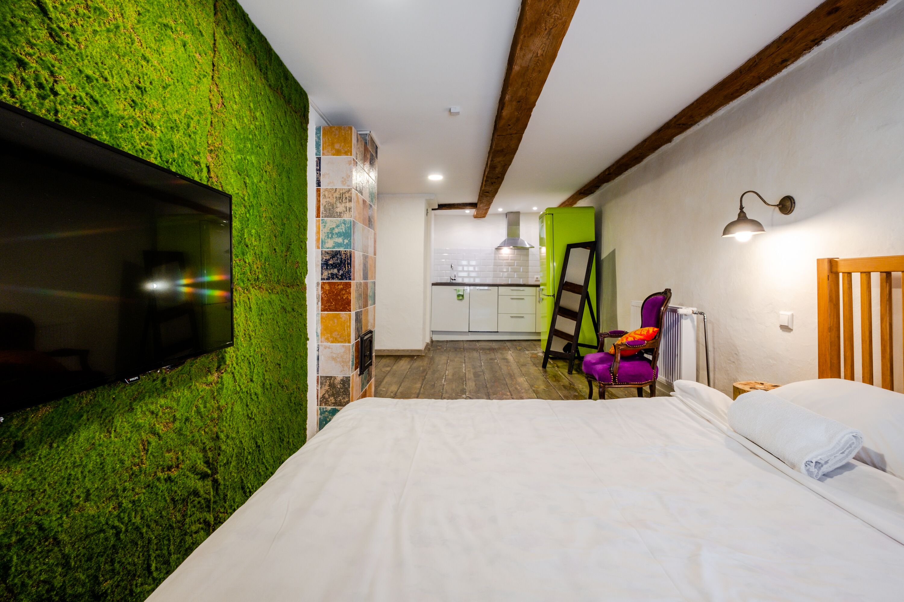 A bedroom with a green moss wall, a flat-screen TV, a wooden ceiling beam, a bed with white linens, a purple chair, and a kitchen in the background.