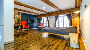 A bedroom with a wooden floor, a bed, a desk with a chair, a TV, and a window with curtains.
