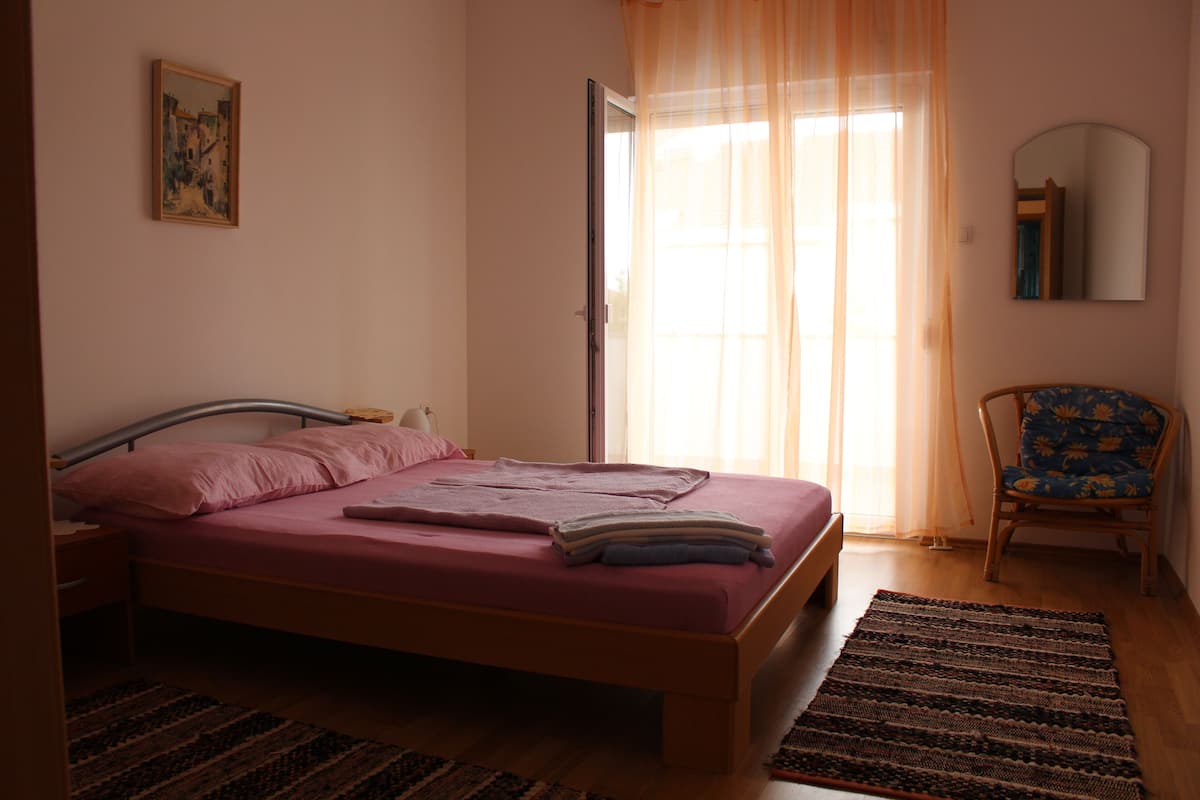 Une chambre à coucher avec un lit, une chaise, une fenêtre avec des rideaux, un tableau au mur et un miroir.