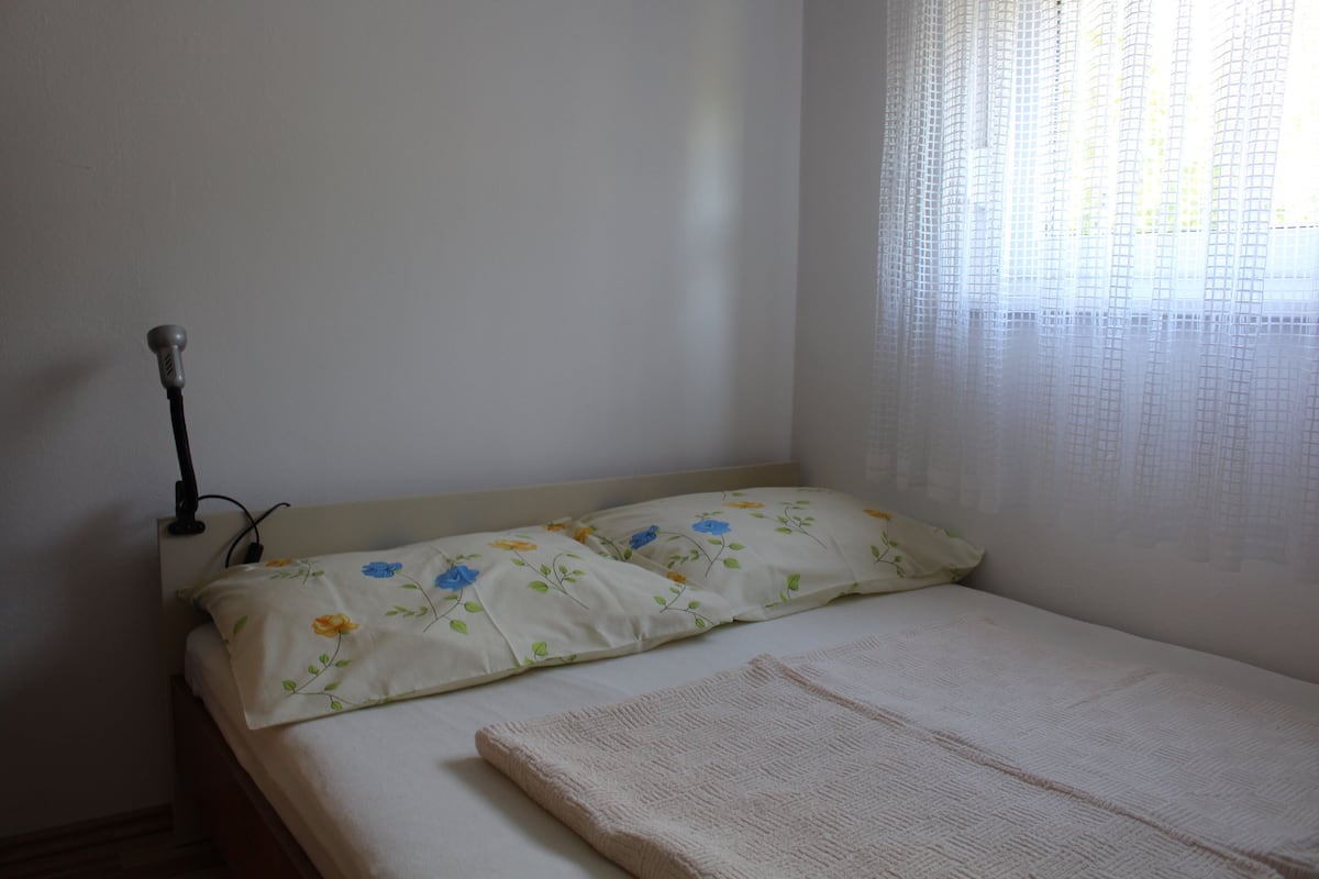 Un lit simple avec une literie à motifs floraux, une lampe de chevet et une fenêtre ornée de rideaux légers.