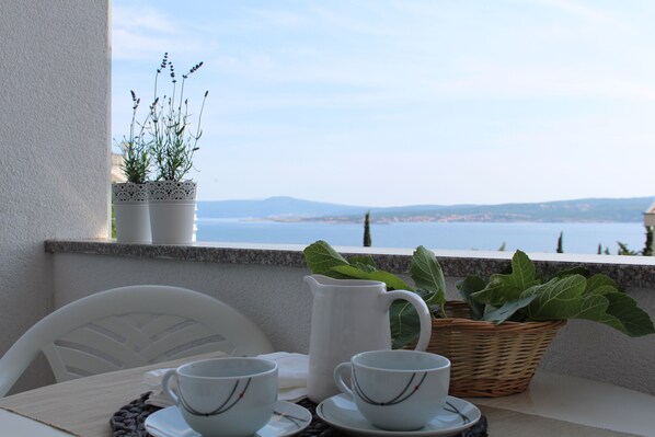 Un balcon avec une table dressée pour deux personnes, une carafe, des verres et un panier rempli de feuilles vertes.