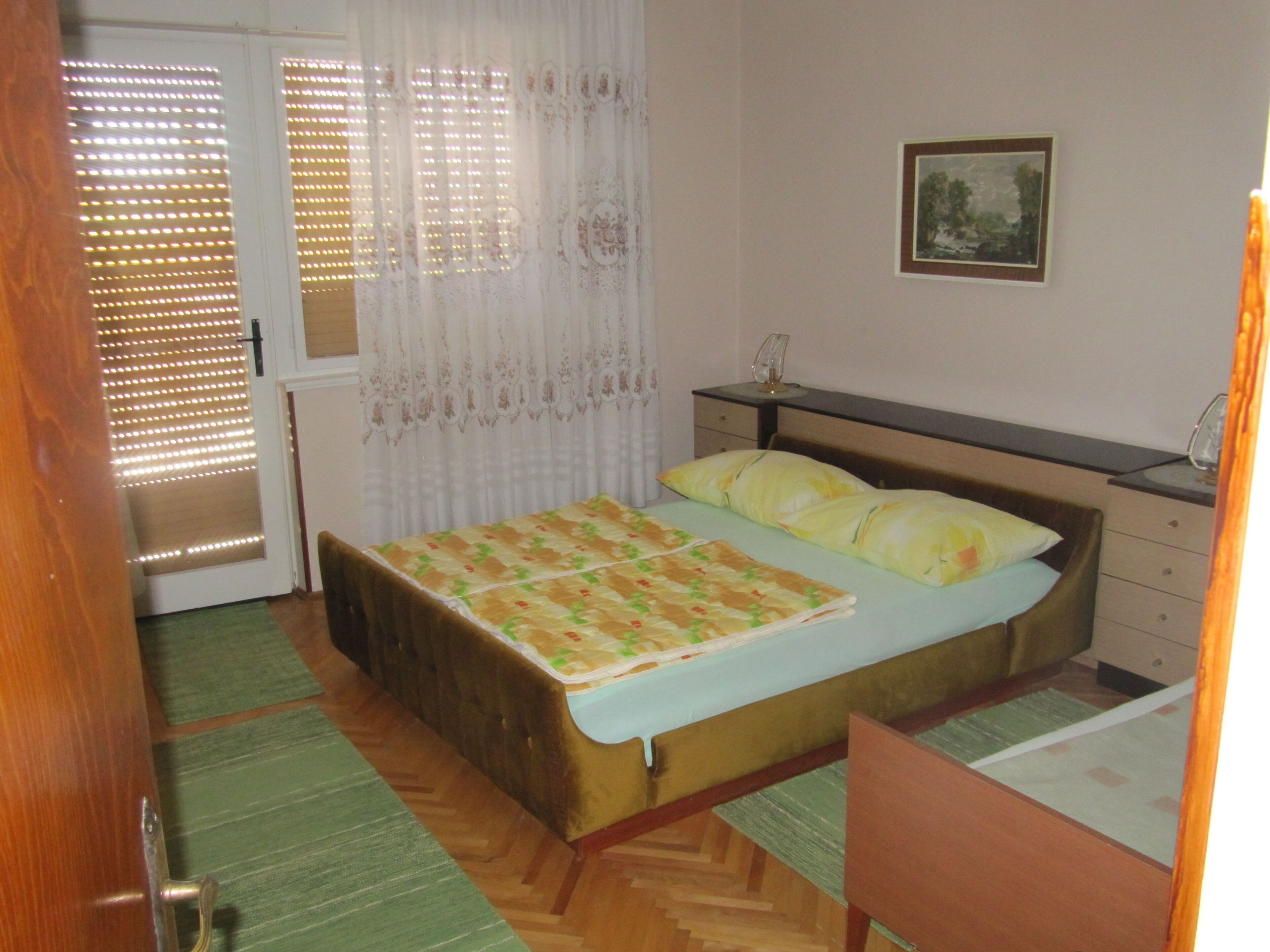 A bedroom with a bed, a bedside table, a chair, a window with blinds, and a picture on the wall.