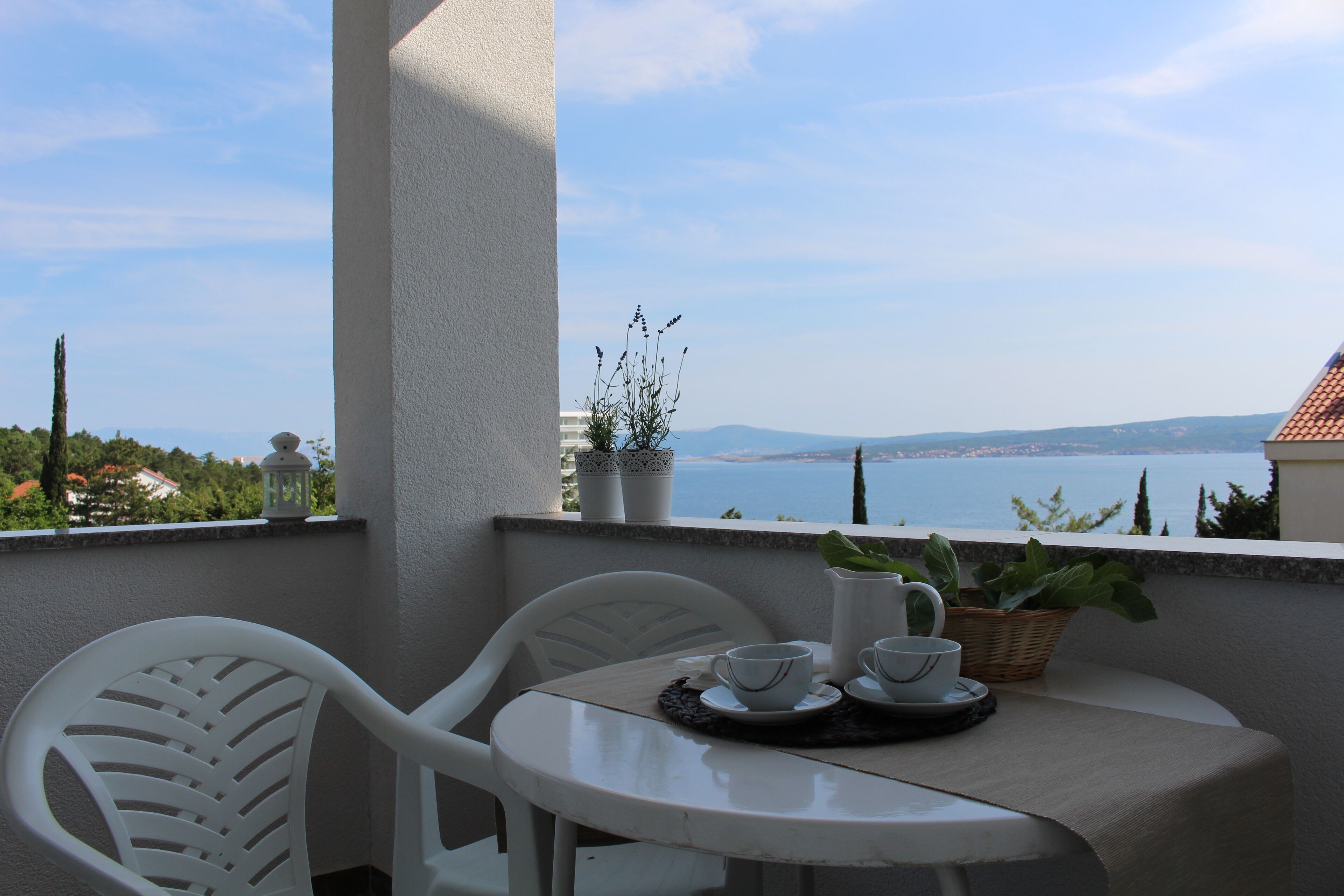 A balcony with a table set for two, chairs, a potted plant, and a view of the sea.