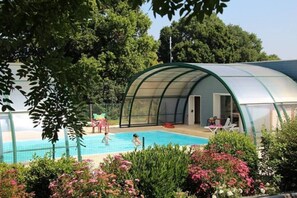 A covered swimming pool with a curved roof, surrounded by lush greenery and flowering plants. - Camping Les Rulières (Saint-Valérien)