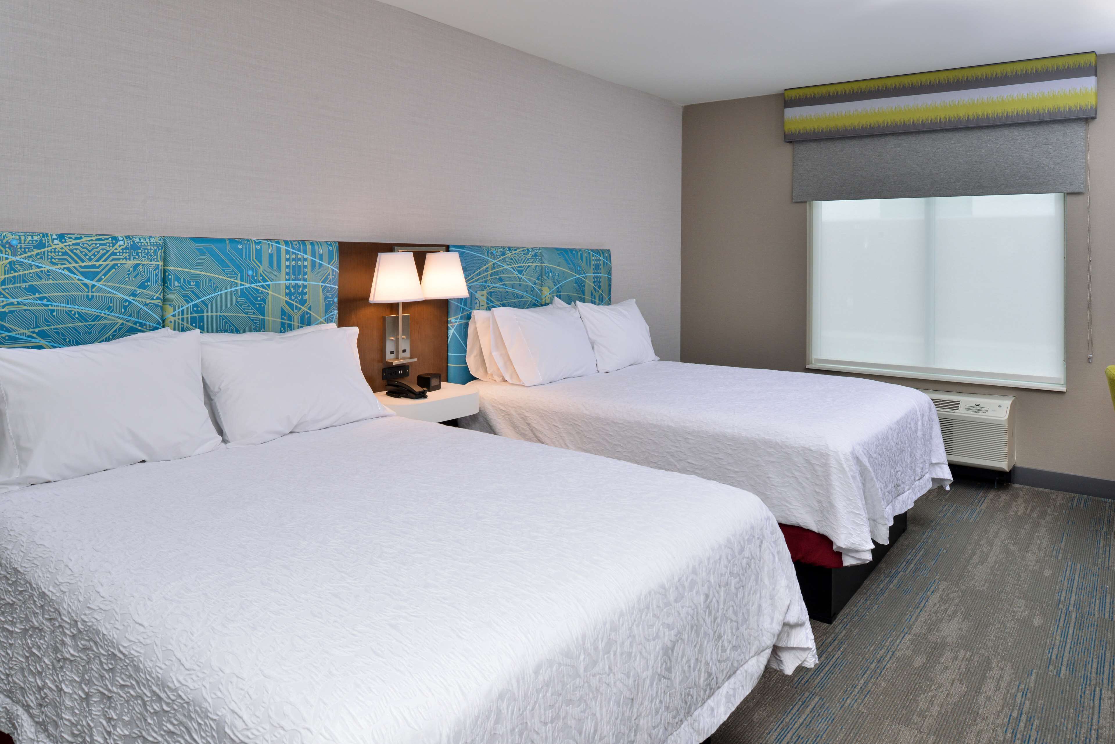 A hotel room with two beds, a nightstand with a lamp, a large window with a view, and a wall with a blue abstract design.