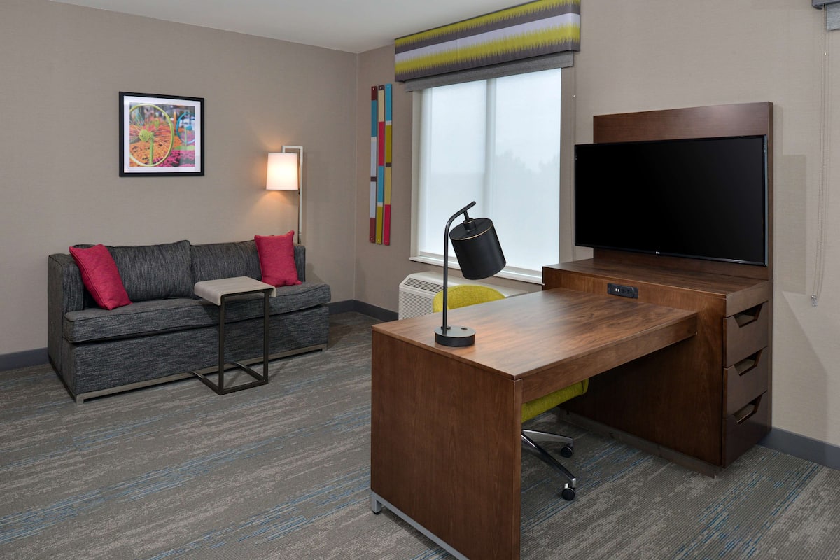 A modern hotel room with a grey sofa, a wooden desk, a flat-screen TV, and a colorful abstract painting on the wall.