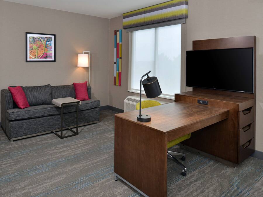 A modern hotel room with a grey sofa, a wooden desk, a flat-screen TV, and a colorful abstract painting on the wall.