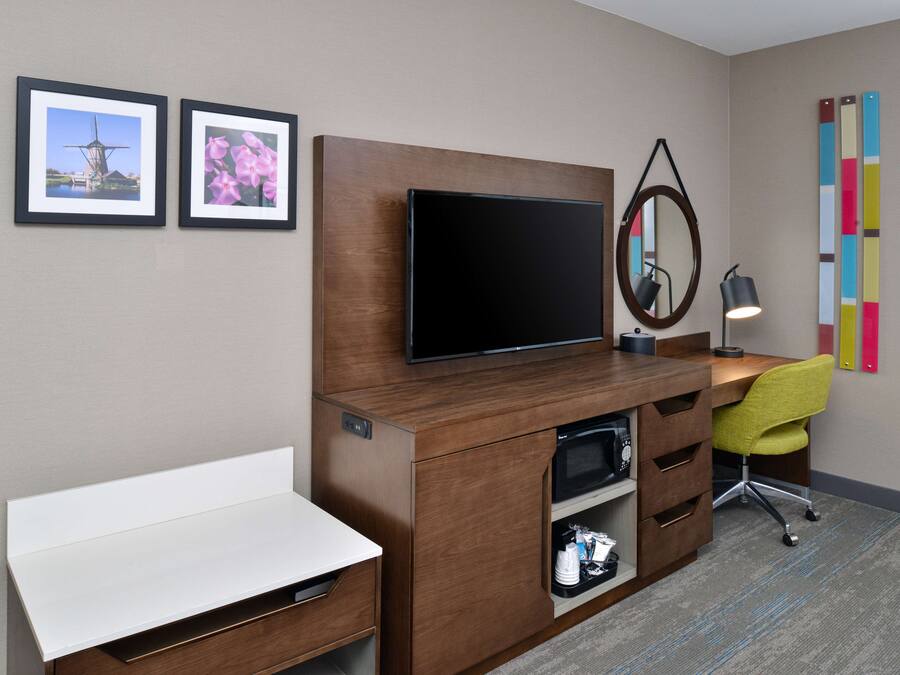 Habitación de hotel con escritorio de madera, televisor de pantalla plana, espejo, silla y dos cuadros en la pared.
