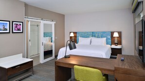 A hotel room with a bed, bedside tables, a desk, a chair, a shower area, and artwork on the walls.
