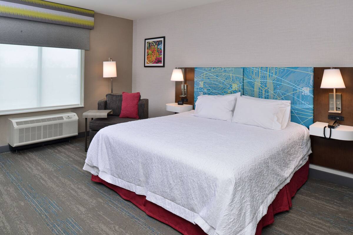 A hotel room with a large bed, two bedside tables with lamps, a window with a view, a wall-mounted air conditioner, and a red accent wall.