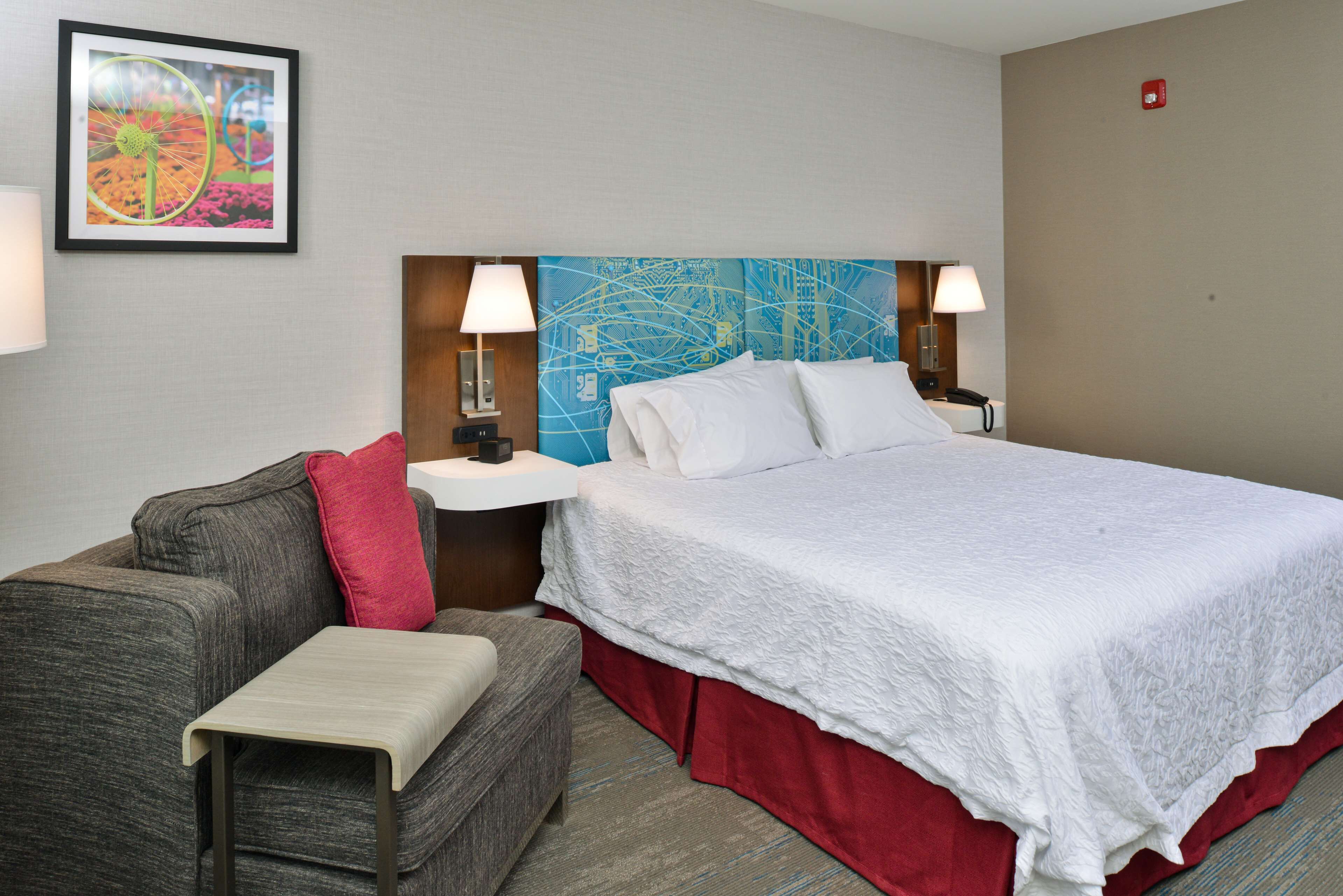 A hotel room with a large bed, a gray sofa, a small table, and a wall-mounted lamp.
