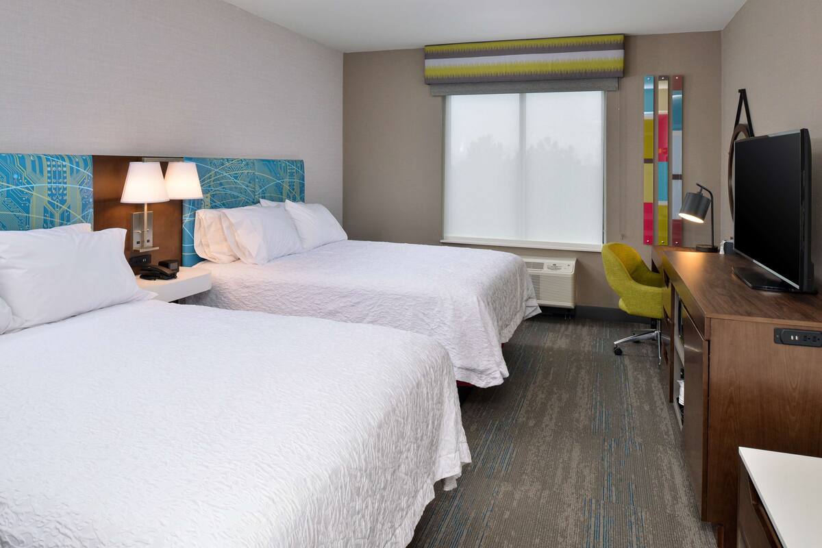 A hotel room with two beds, a desk, a chair, a television, and a window with blinds.