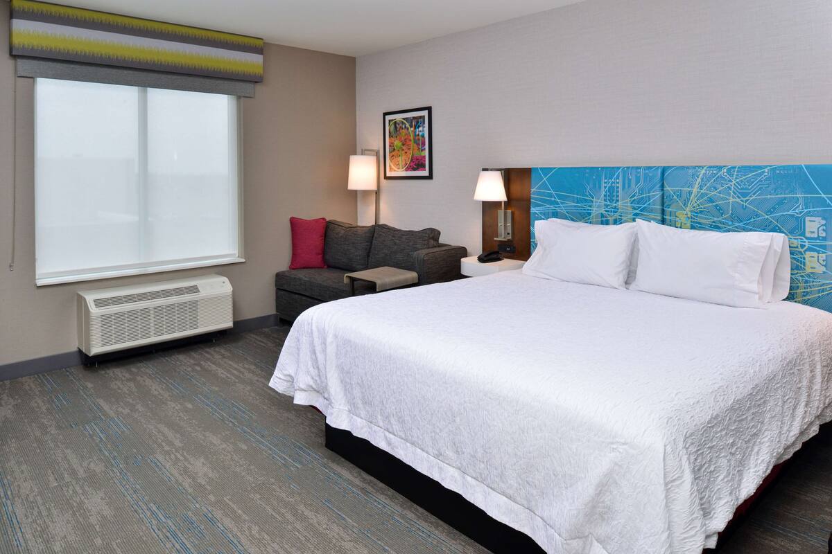 A hotel room with a large bed, a desk, a chair, a heater, a window with blinds, and a wall with a map design.