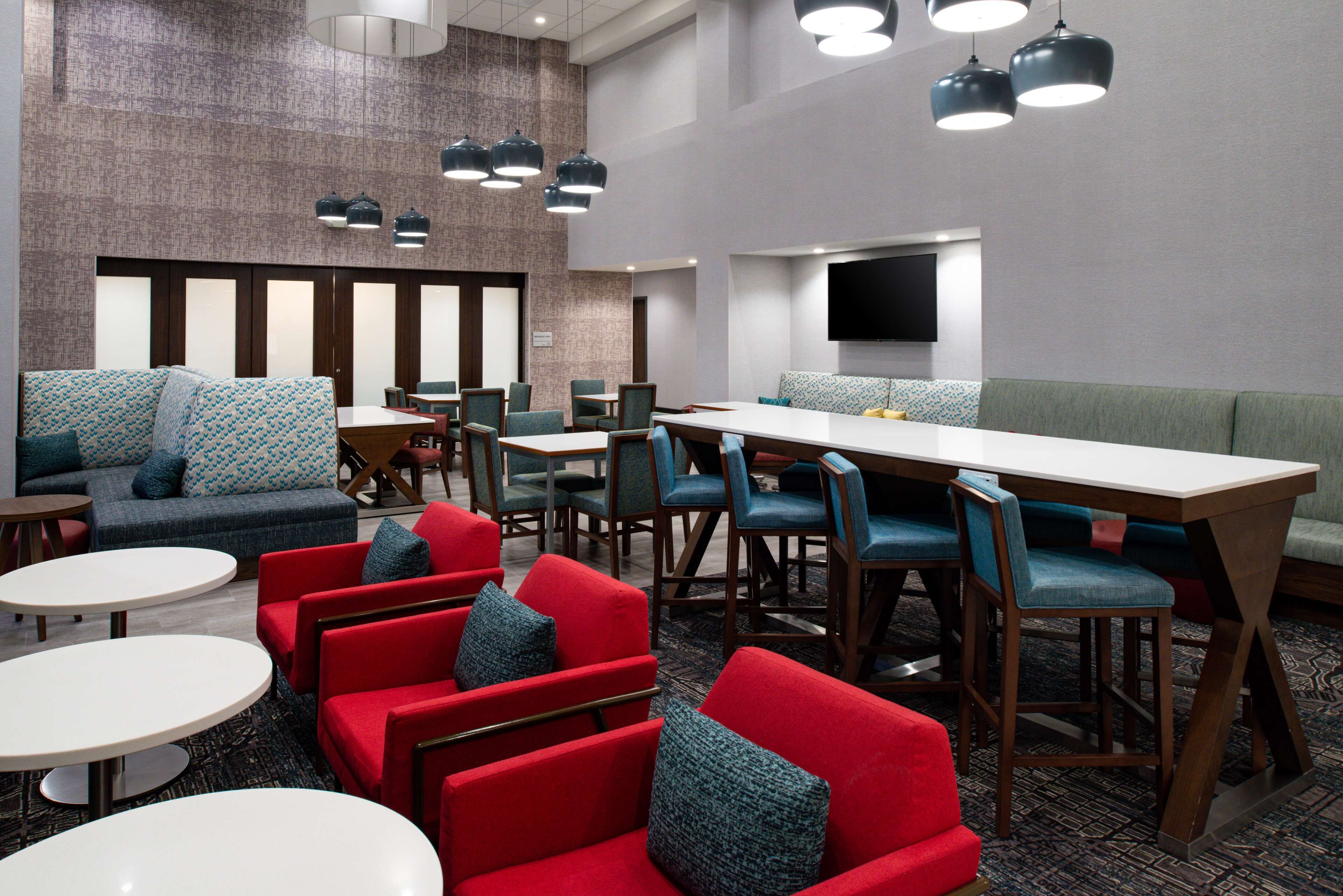 A modern hotel lobby with a mix of seating options including red and blue upholstered chairs, a long communal table, and a patterned sofa.