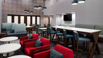 A modern hotel lobby with a mix of seating options including red and blue upholstered chairs, a long communal table, and a patterned sofa.