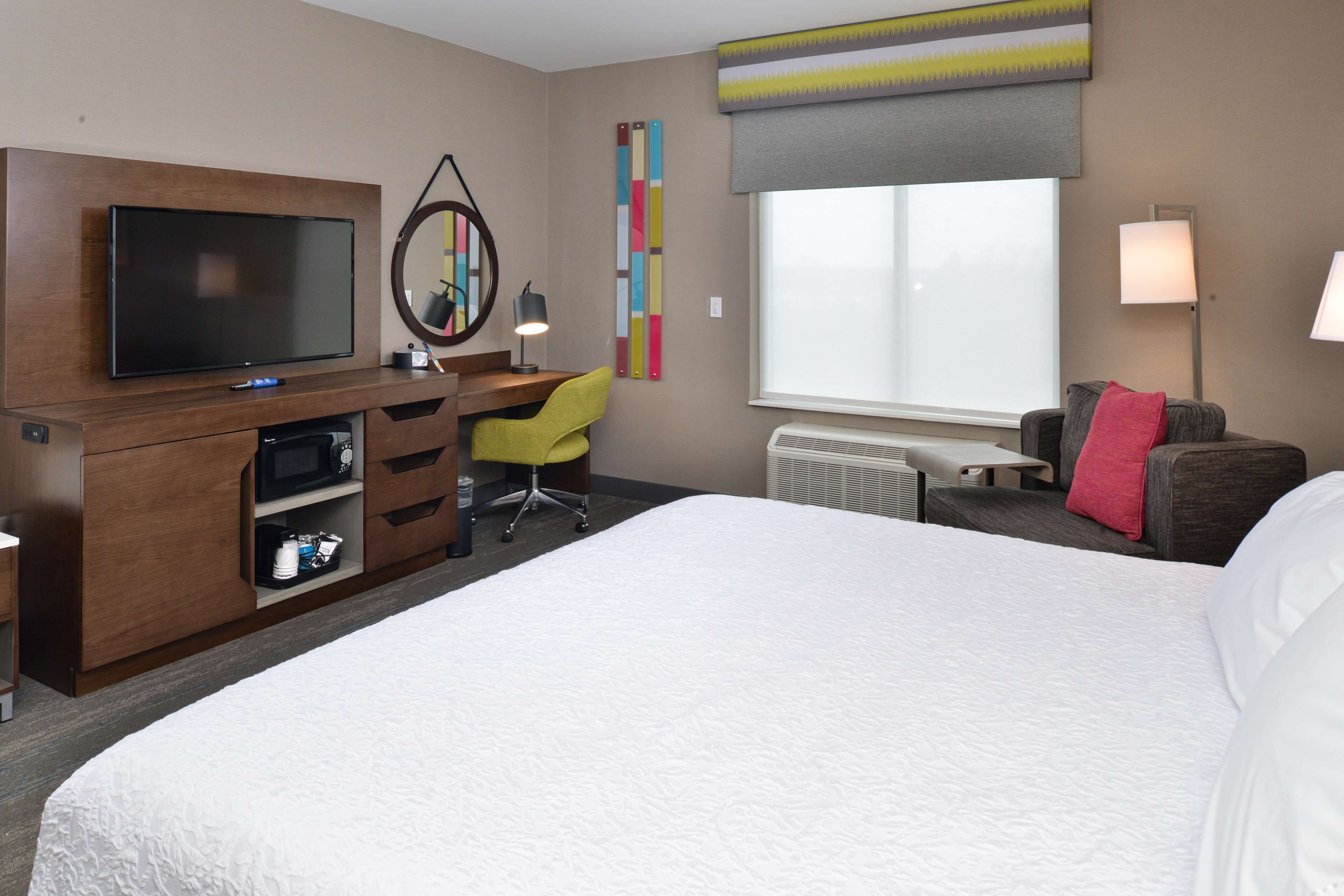 A hotel room with a bed, a desk with a TV, a chair, a lamp, and a window.