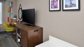 A hotel room with a wooden desk, a television, a yellow chair, a white bed, and two framed pictures on the wall.