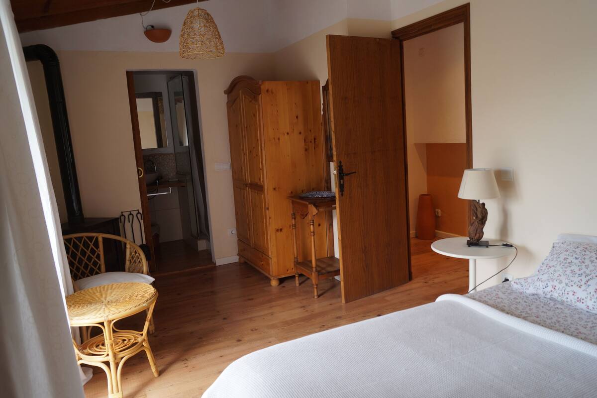 Un dormitorio con una cama, una silla de mimbre, un armario de madera, una mesita con una lámpara y una puerta que da a otra habitación.