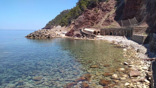 A clear, rocky shoreline with a small building structure on a hillside. - Sa Plana Petit Hotel (Estellencs)