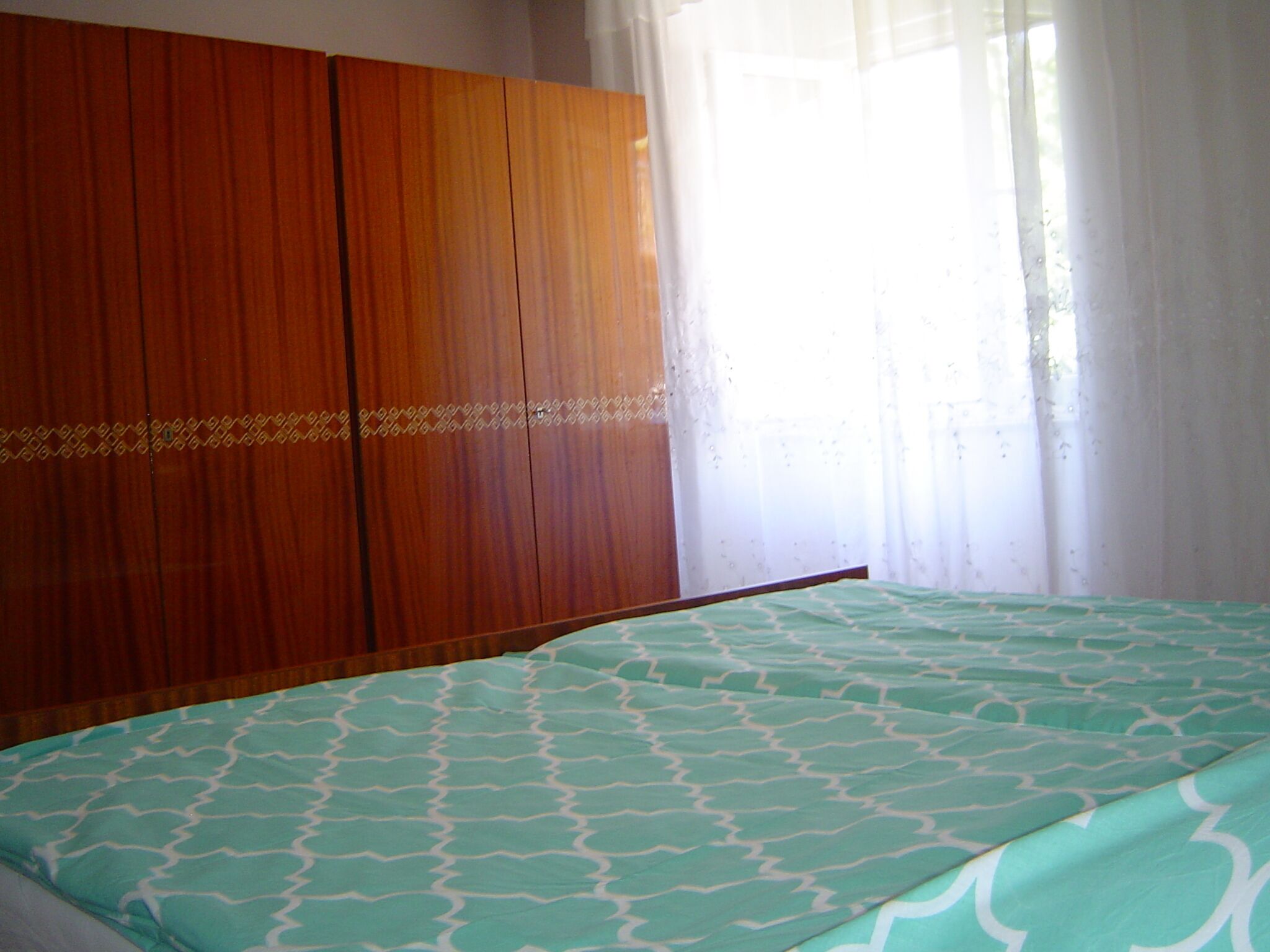 Ein Schlafzimmer mit einem Bett, einem Holzschrank und einem Fenster mit durchsichtigen Vorhängen.