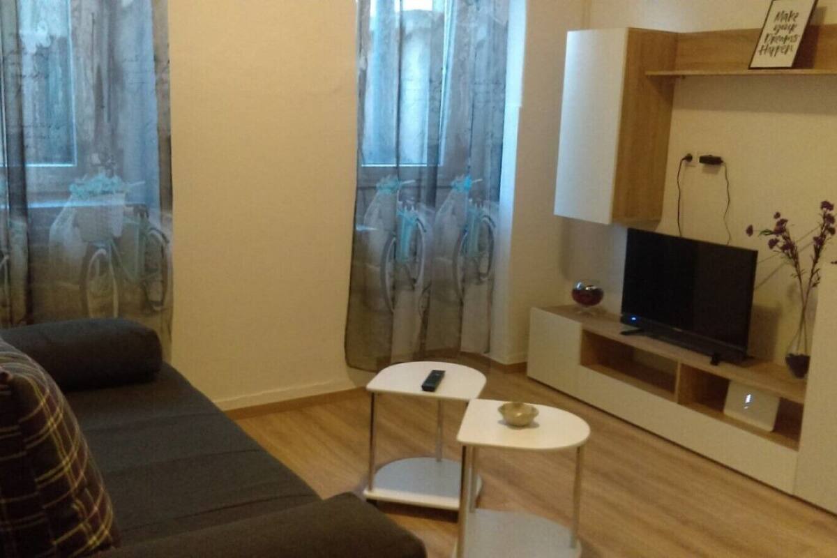 A compact living space with a sofa, a small white table, a TV on a wooden stand, and a window with sheer curtains.