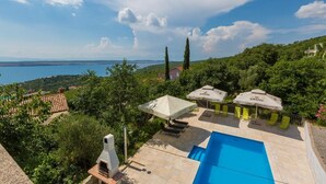 A pool area with lounge chairs and umbrellas, surrounded by greenery and overlooking a body of water. - Apartments Silly (Crikvenica)