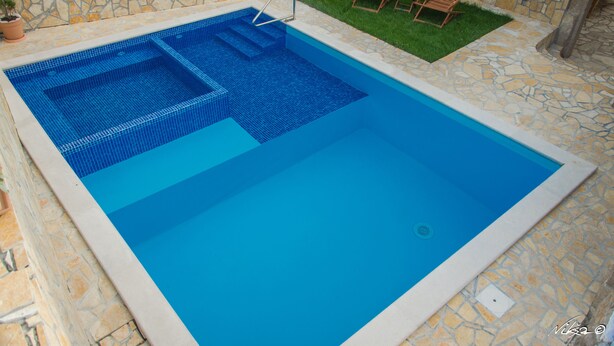 A swimming pool with a stone-tiled edge, a small grassy area with two wooden chairs, and a stone wall in the background. - Apartment Milly (Vinodol)