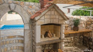A stone-built outdoor oven with a brick chimney, a stone archway, and a pool in the background. - Apartment Milly (Vinodol)