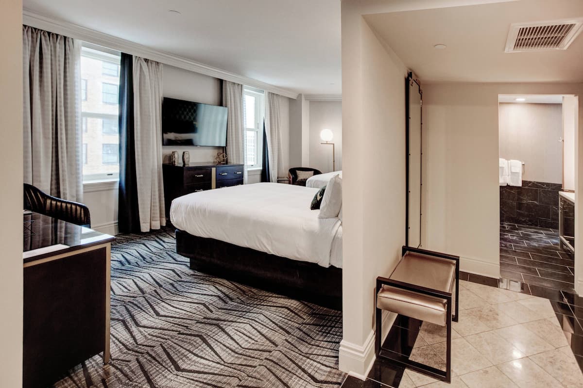 A hotel room with a large bed, a desk, a chair, a TV, and a bathroom visible through an open door.