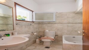 Un baño con lavamanos, inodoro y bañera, paredes con azulejos y una ventana con vistas.