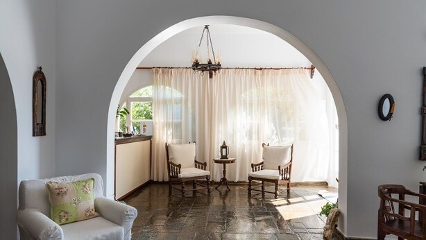 Una habitación con suelo de piedra, un sillón blanco, dos sillas de madera, una mesita y una araña.