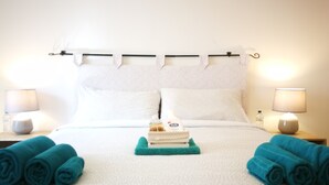 Una cama bien hecha con sábanas blancas, una mesita con una botella de loción y dos toallas enrolladas sobre la cama.
