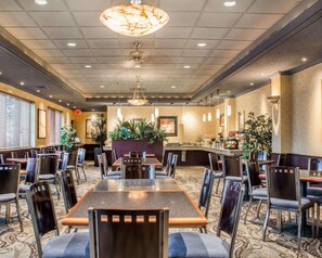 A spacious dining area with multiple tables and chairs, a central bar area, and decorative plants. - Quality Inn & Suites Riverfront (Oswego)