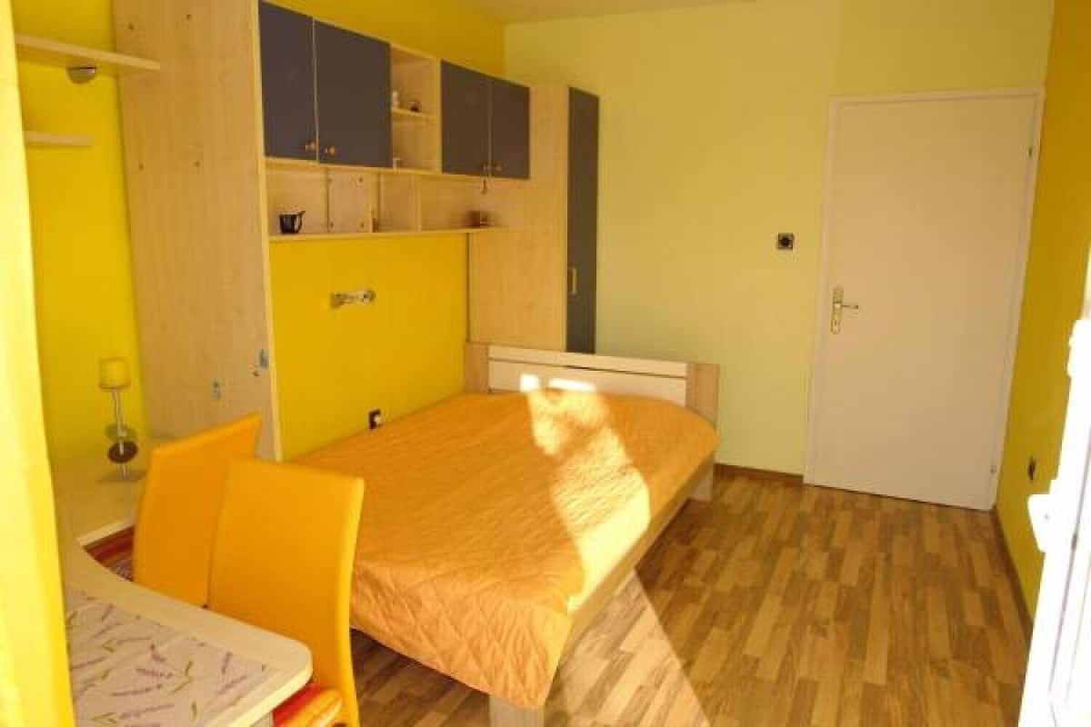 A small, brightly lit bedroom with a single bed, a chair, and a desk.