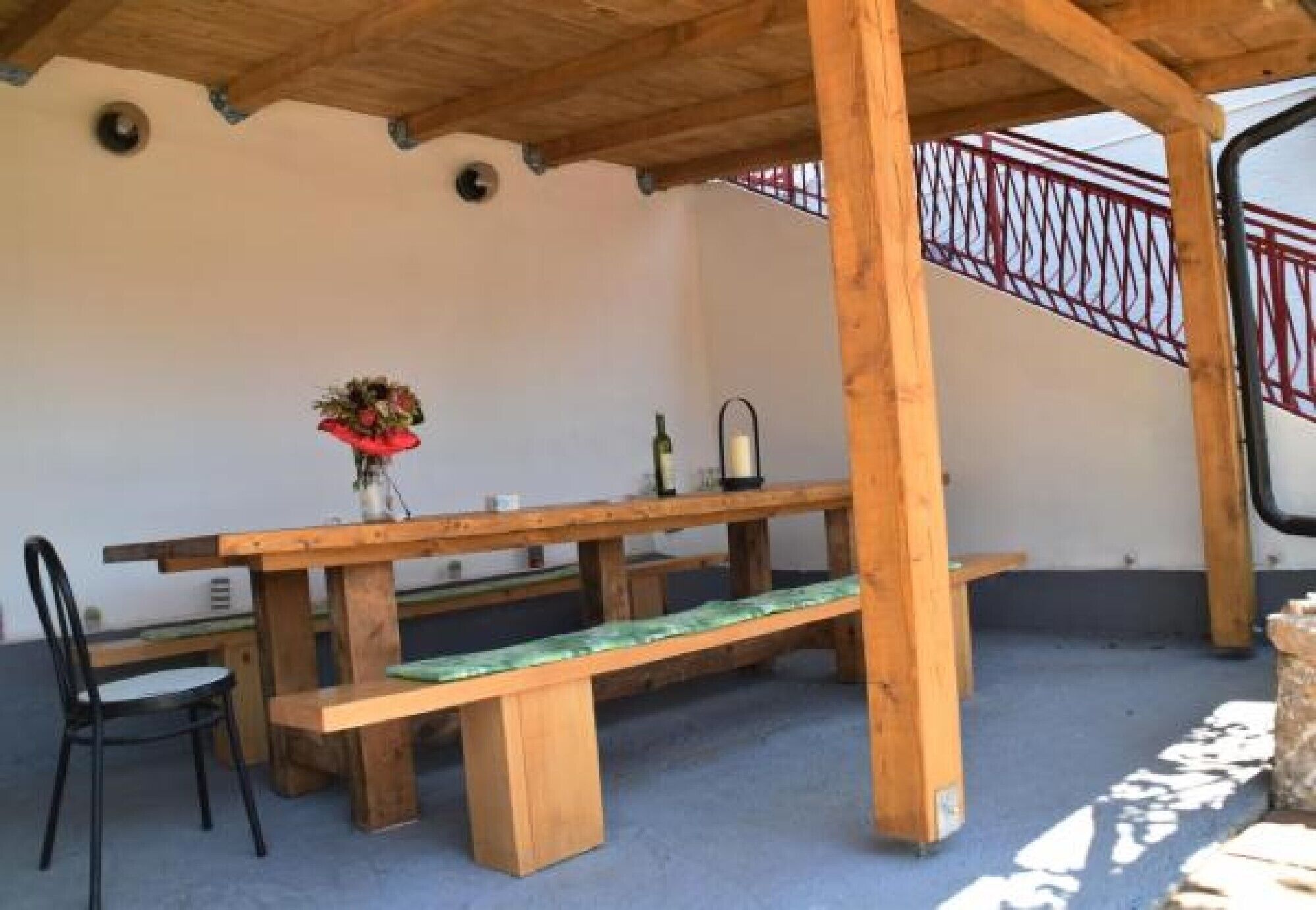 A wooden table with benches under a wooden pergola, a chair, and a railing in the background.
