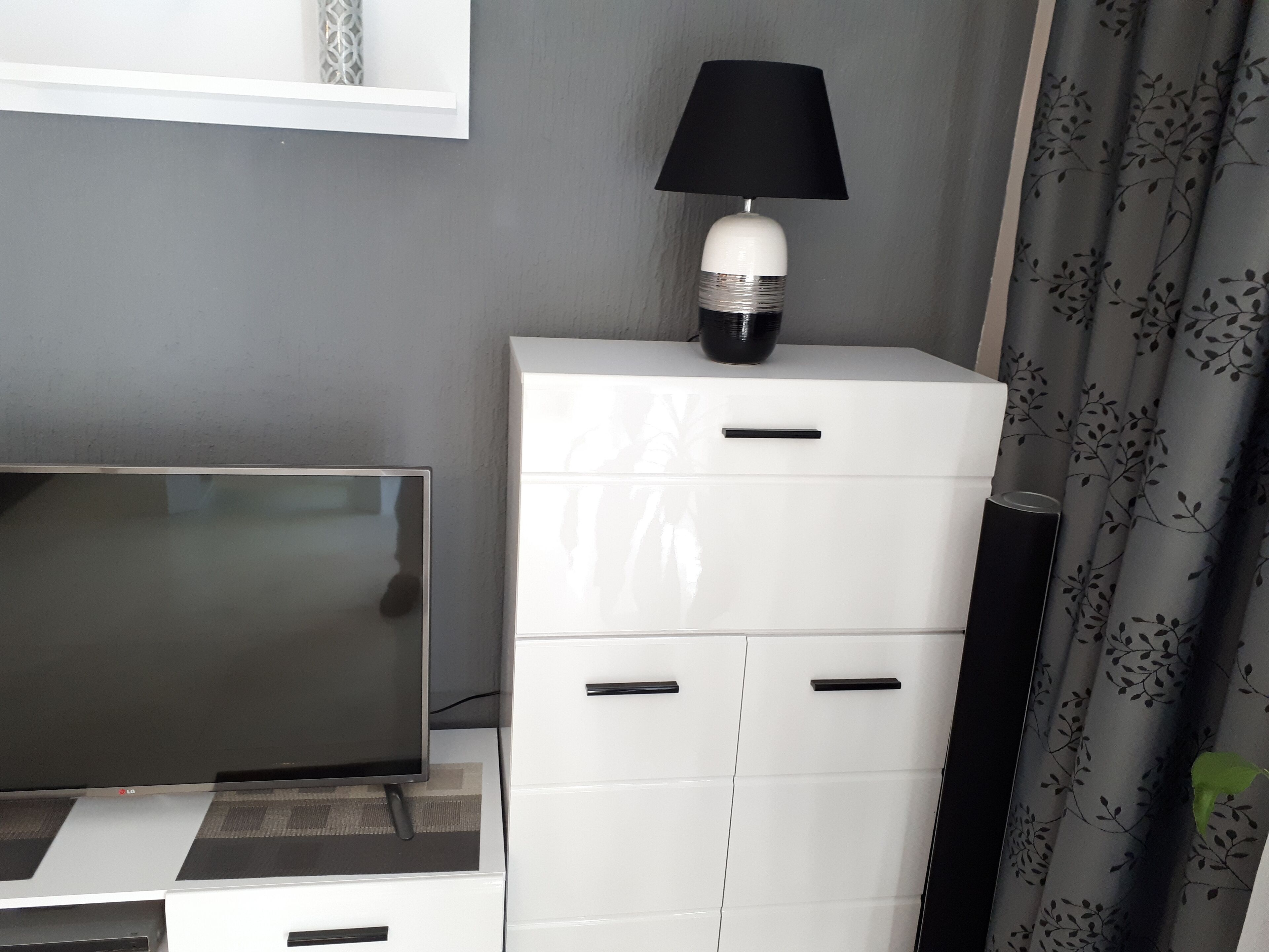 Une chambre moderne dotée d’un téléviseur à écran plat, d’une commode blanche avec des tiroirs, d’une lampe noire et d’un rideau à motifs.
