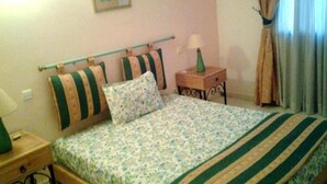 Quarto com cama, mesa de cabeceira, abajur, quadro na parede e cortinas na janela.
