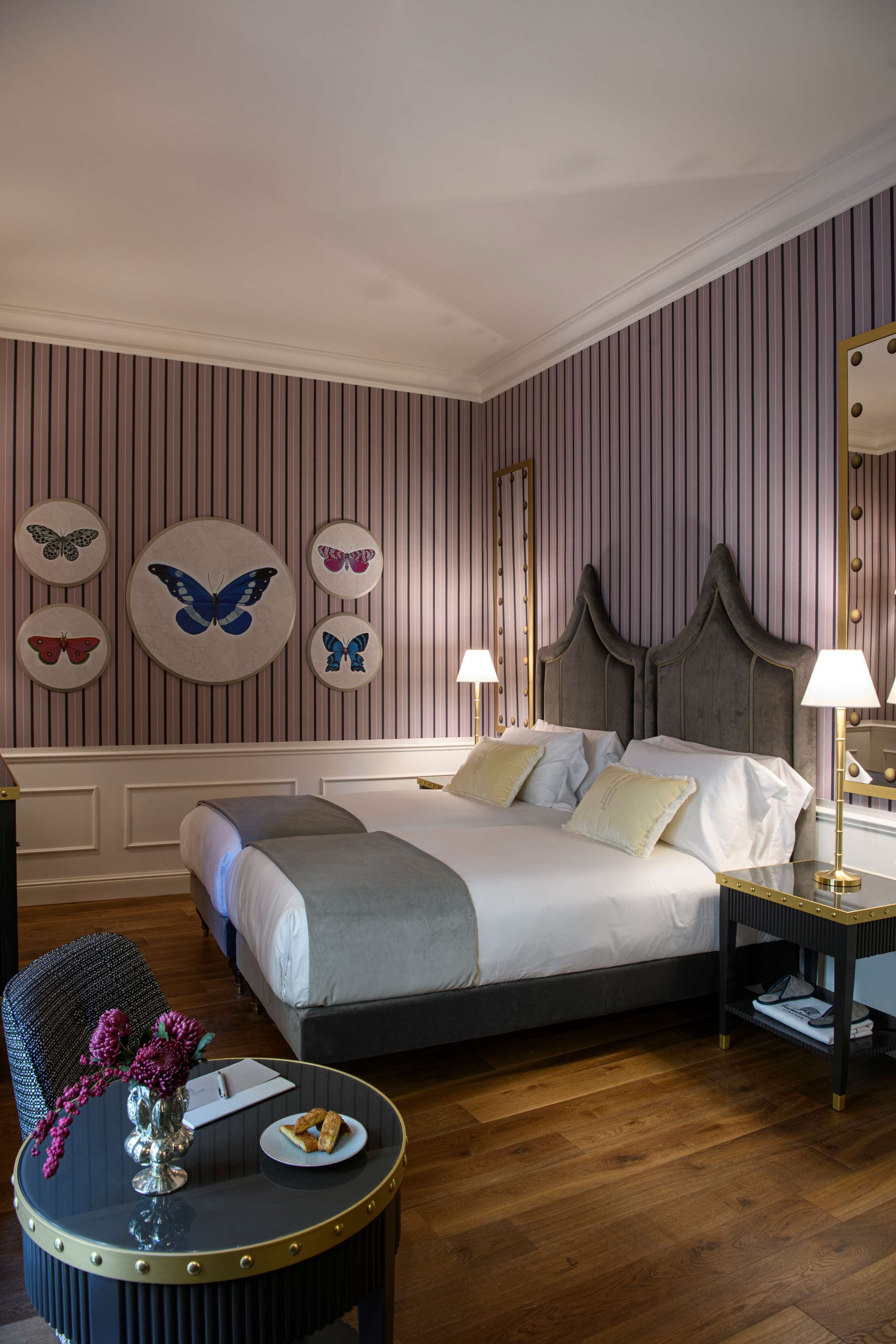 Una habitación de hotel con cama, mesitas de noche, una mesa redonda con un florero y un plato de comida, y decoración de mariposas en la pared.