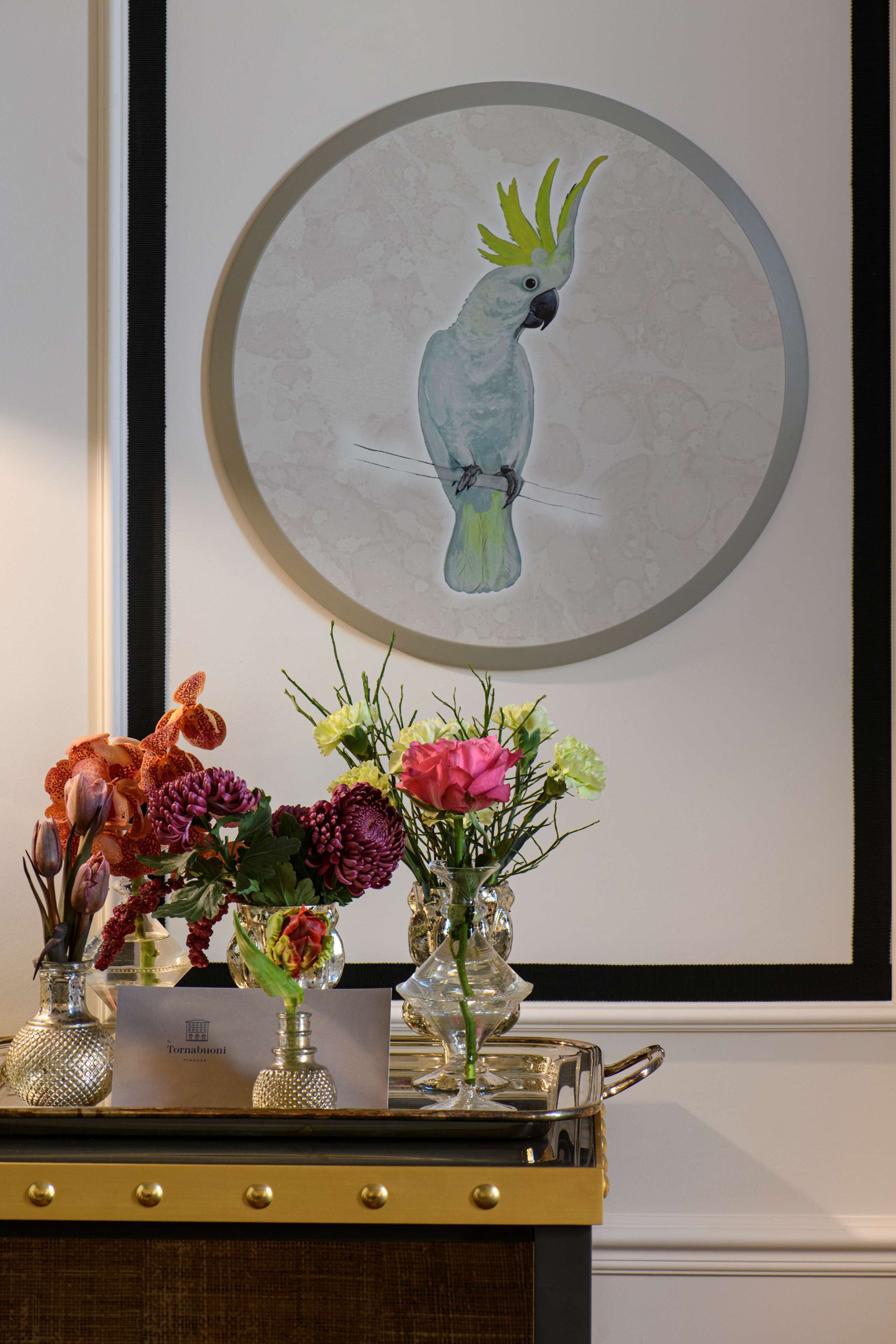 Un'opera d'arte incorniciata raffigura un pappagallo, un tavolo con vasi di fiori e un cartello con su scritto "Tornatiomi".