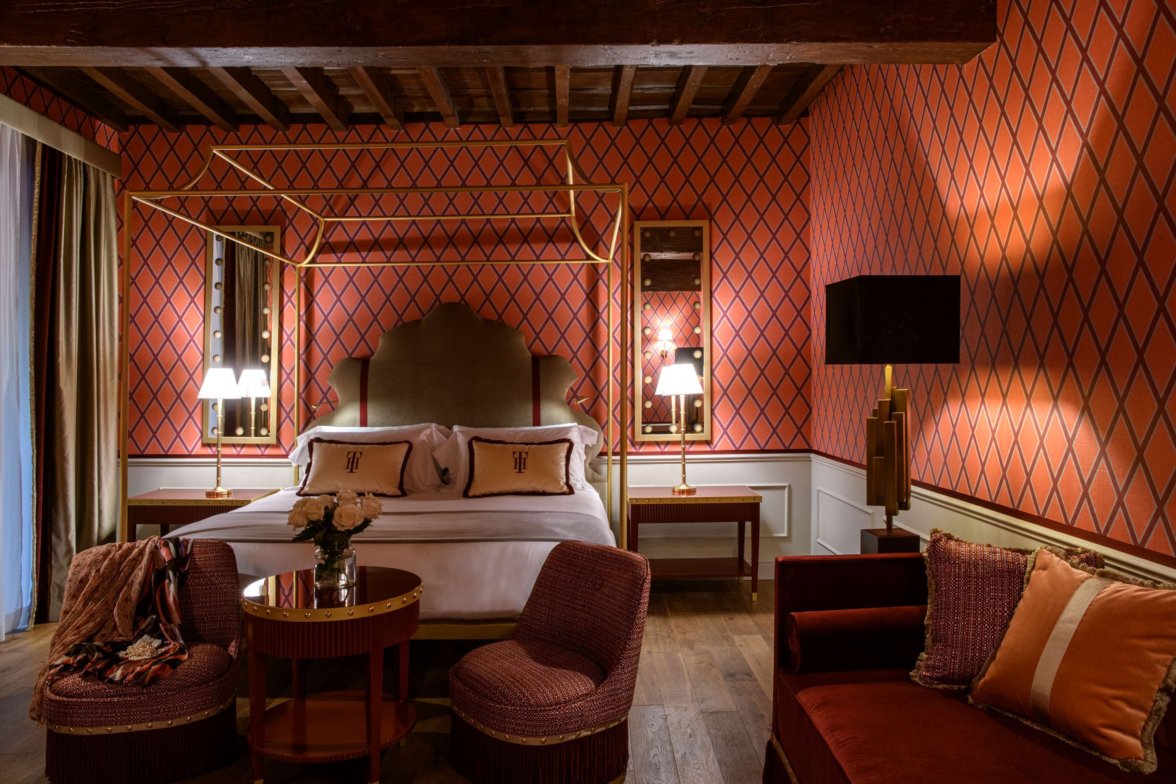A hotel room with a large bed, a sitting area with a sofa and armchairs, a small table with a vase of flowers, and a large mirror above the bed.