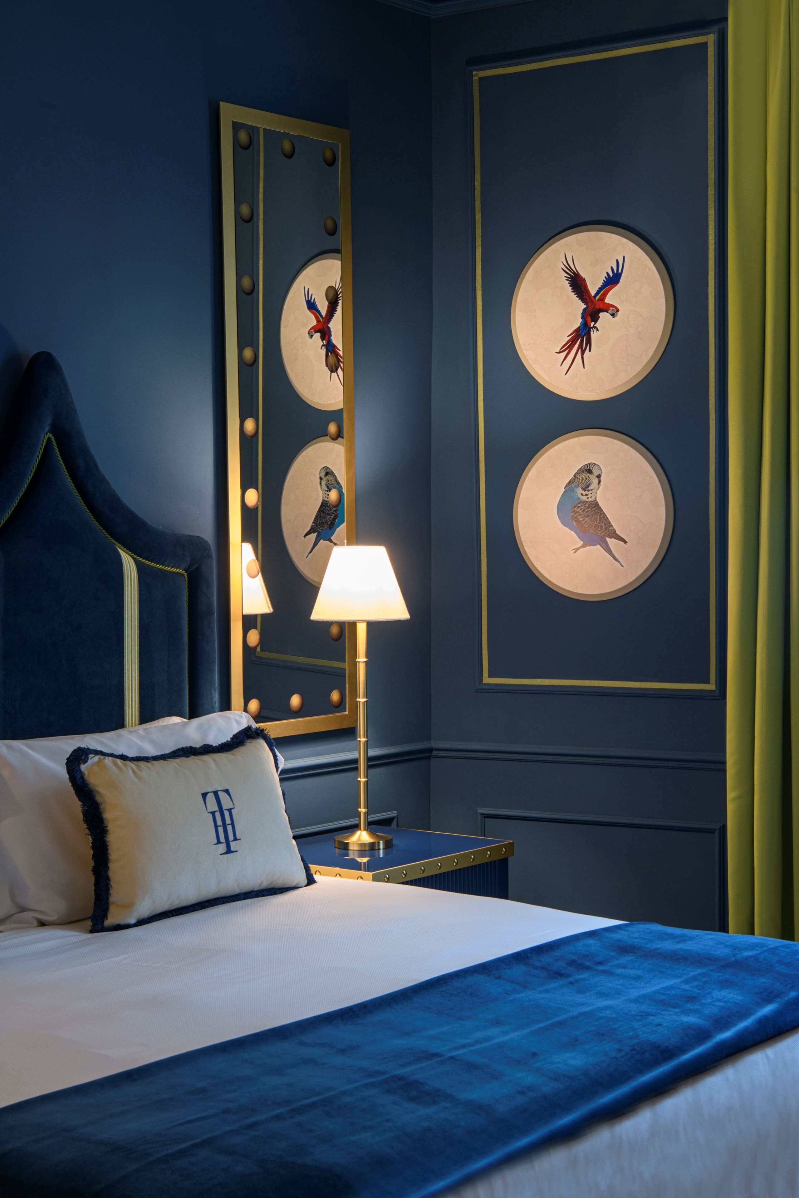 Un lit bien fait, recouvert d’une couvre-lit bleue et agrémenté d’un oreiller orné d’un monogramme. Un miroir rond, décoré d’un motif d’oiseau, est accroché au mur, ainsi qu’une lampe posée sur une table de chevet et un tableau encadré représentant un oiseau.