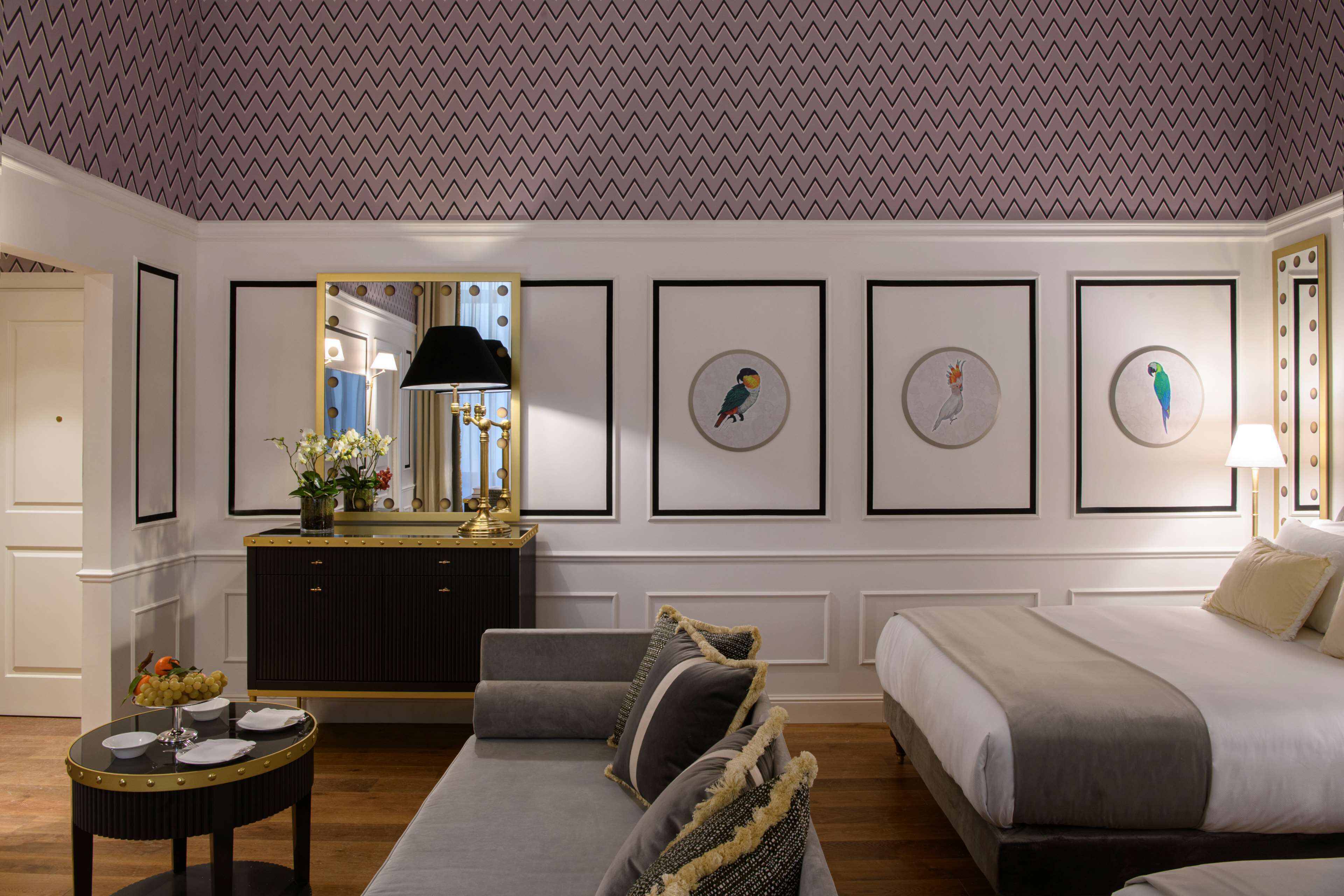 A hotel room with a bed, a sofa, a small table with a vase of flowers, a mirror, and framed artwork on the wall.