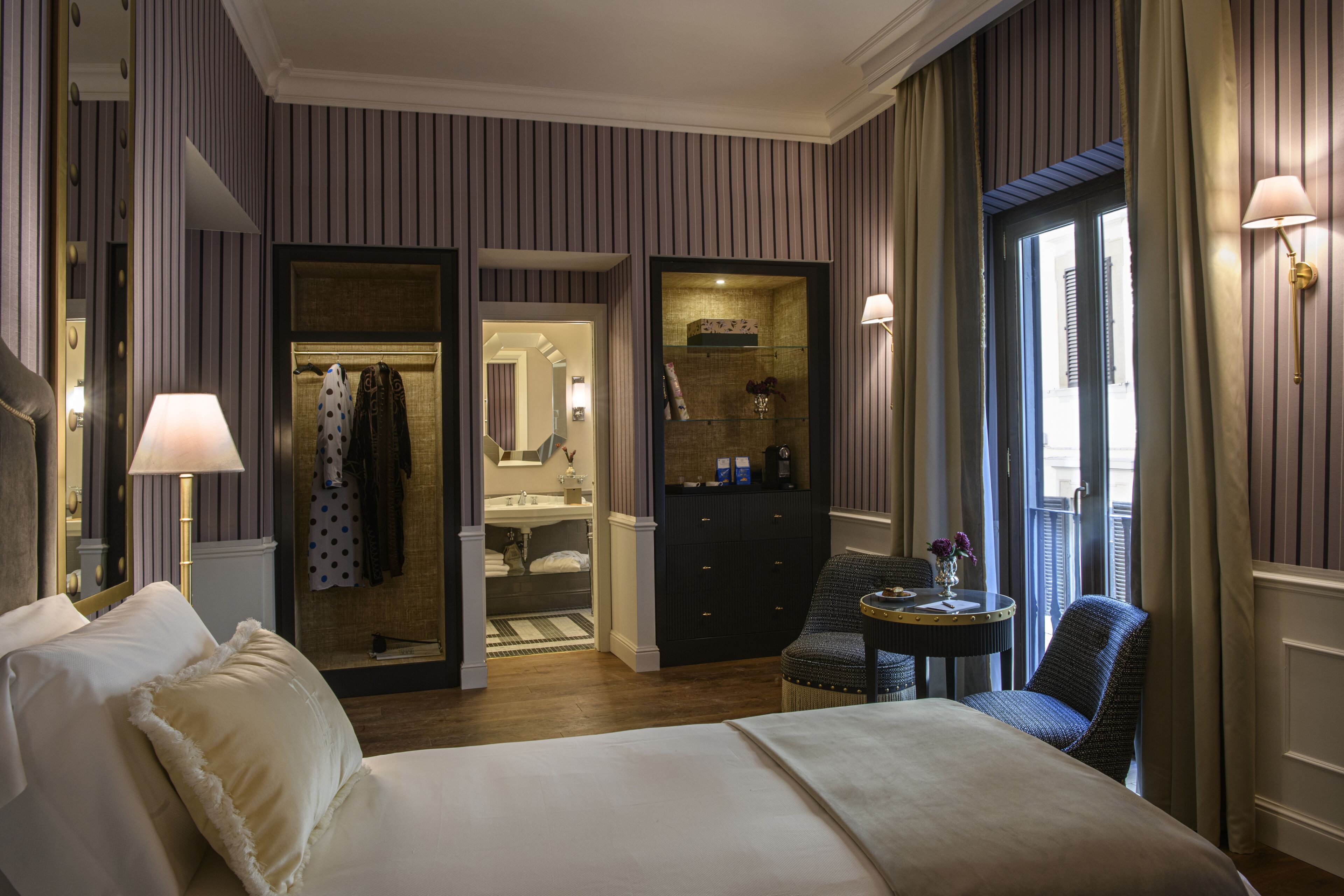 A hotel room with a large bed, a sitting area with a chair and table, a vanity with a mirror, and a walk-in closet.