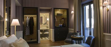 A hotel room with a large bed, a sitting area with a chair and table, a vanity with a mirror, and a walk-in closet.